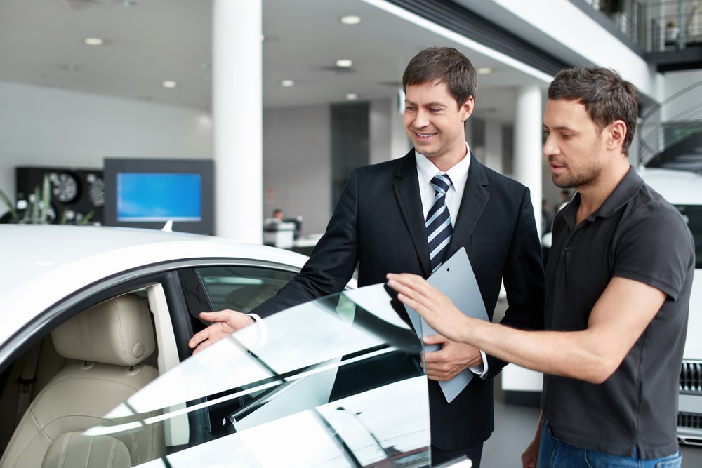 Two men in a car dealership, one in a suit holding a tablet, discussing a car.