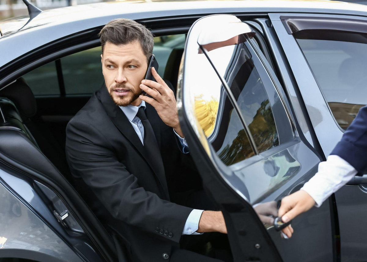 Man in a suit sitting in a car, talking on a phone with a driver opening the door.